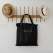 Black tote bag with 'HEALIX' logo hanging on a wooden rack with hats on a white wall.