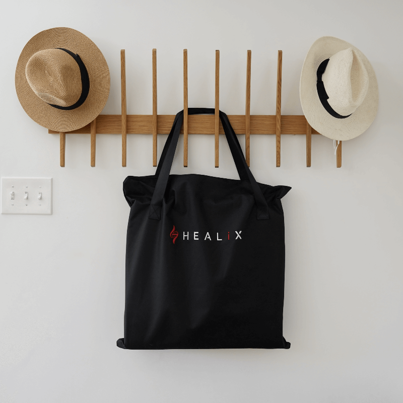 Black tote bag with 'HEALIX' logo hanging on a wooden rack with hats on a white wall.