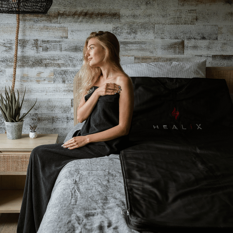 Woman sitting on a bed with a black sauna blanket featuring 'HEALIX' branding.