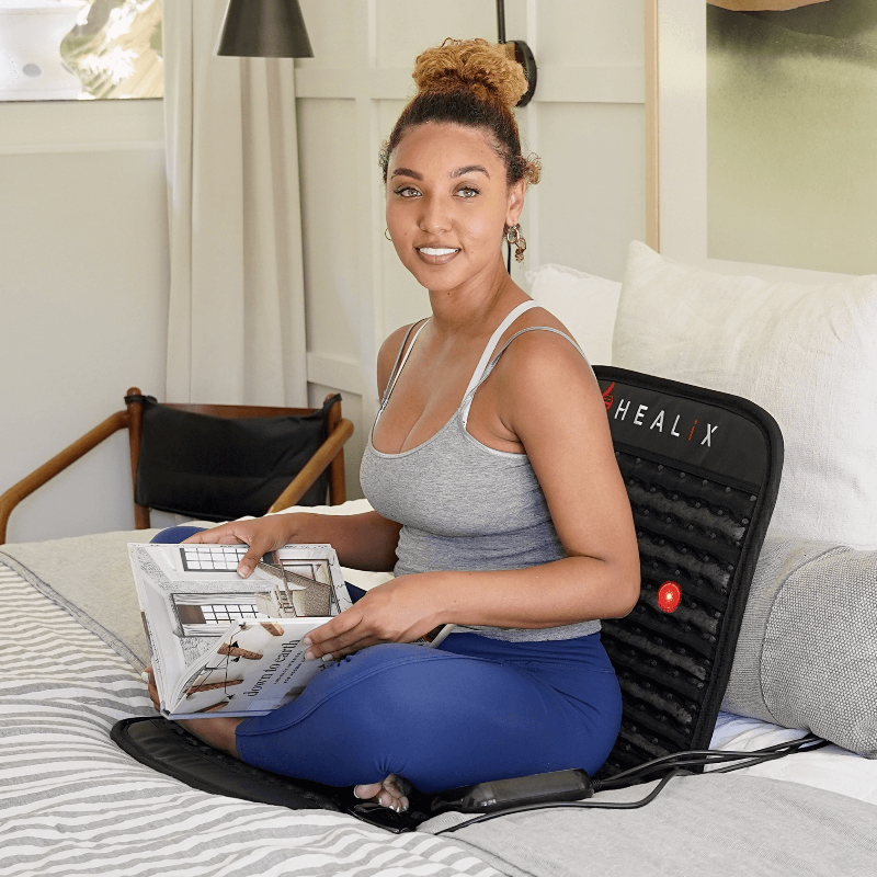 Woman using a HEALIX red light mat on a bed while reading a magazine.