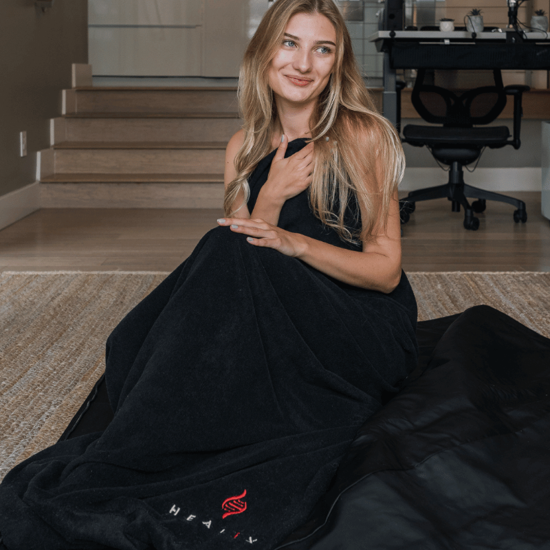Woman wrapped in a black sauna blanket with a visible brand logo in a home setting