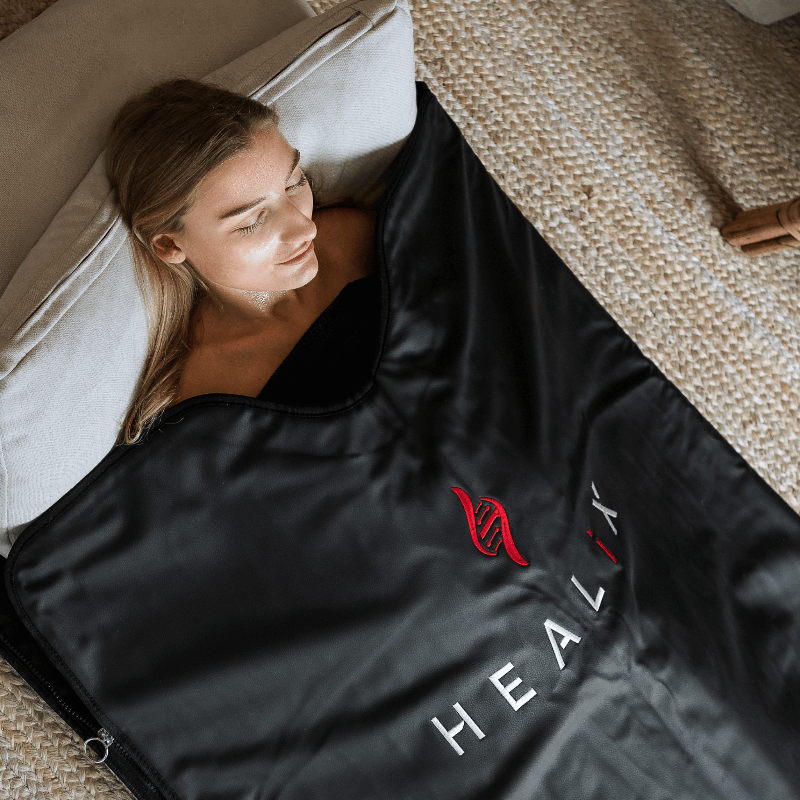 Woman lying under a black infrared sauna blanket with 'HEALIX' branding.