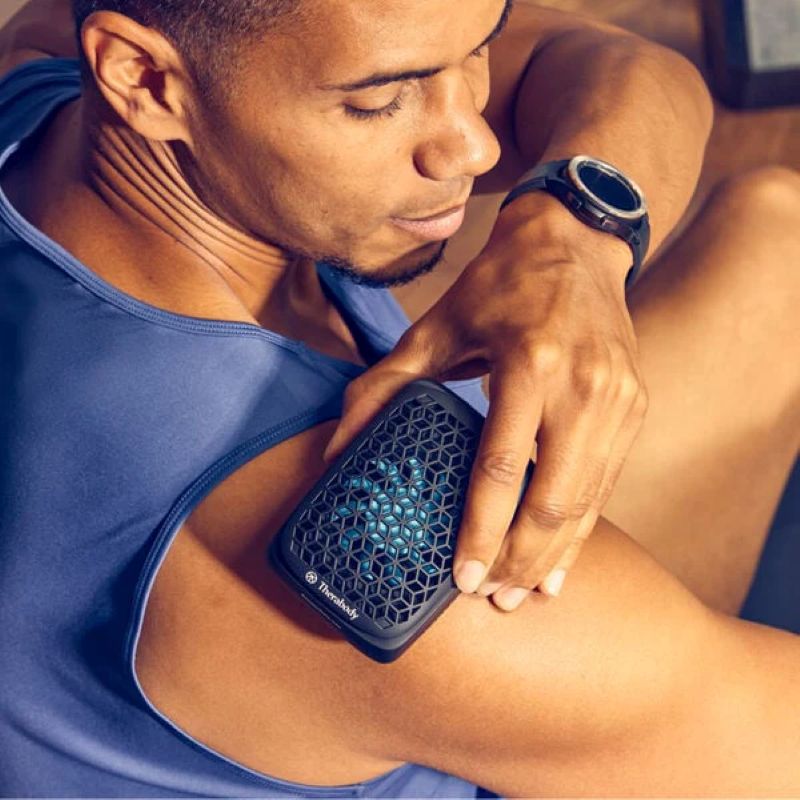 Man using a handheld massager on his arm, wearing a blue tank top.
