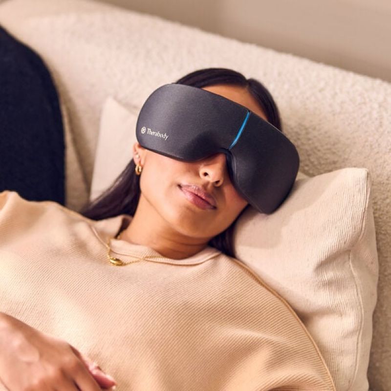 Person wearing a black sleep mask with 'Therabody' branding, lying on a beige pillow.